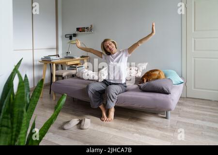 Happy woman waking up alone in morning at home, sitting on bed with dog, stretching body muscles Stock Photo