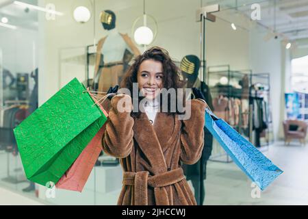 Holiday sale. Beautiful young curly woman in fur coat holding paper colored bags with shopping and gifts for Christmas holidays for family and friends. Shows, brags, rejoices, looks into the camera in a store in a shopping center Stock Photo
