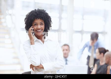 Talking with clients on the phone. An african woman speaking on her cellphone in the office with her colleagues behind her. Stock Photo