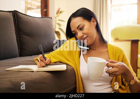 Hispanic woman working remotely sitting on the floor against a sofa drinking coffee in a bright living room. A serious focused young female writing Stock Photo
