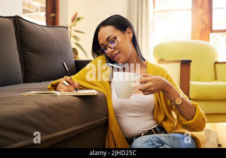 Hispanic woman writing notes sitting on the floor against a sofa drinking coffee in a bright living room. A serious focused young female working Stock Photo