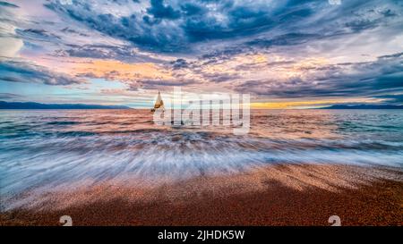 A Sailboat Sails Along The Ocean With A Colorful Cloudscape Overhead Stock Photo