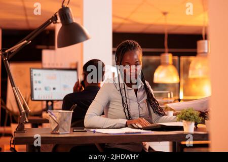 Focused freelancer looking at business charts on documents, analyzing research information on paperwork to create research project. Working with papers and computer in office with big windows. Stock Photo