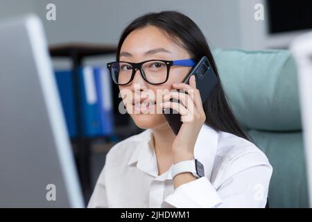 Close-up photo. Portrait of a young beautiful Asian business woman in glasses in a white shirt talking on the phone. He is sitting at the table in the office, holding a hand Stock Photo