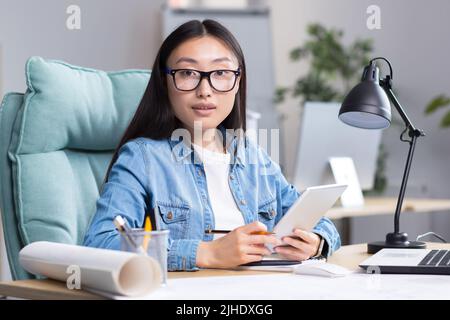 Portrait of a young beautiful Asian architect-designer woman in glasses works on a project, sits at a desk, holds a tablet in her hands. He looks at the camera, smiles. Stock Photo