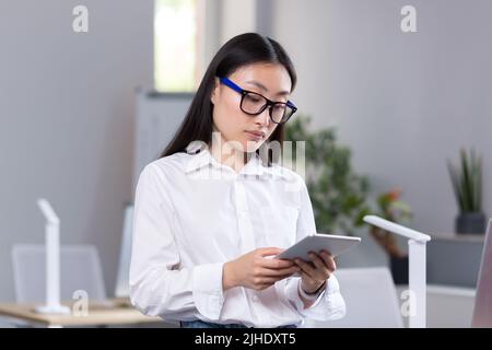 Portrait of young beautiful Asian female student in glasses, white shirt and jeans. Standing in the office, holding a tablet in his hands, studying, typing. Stock Photo