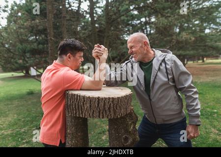 Happy senior father with his adult son with Down syndrome arm wrestling in park. Stock Photo