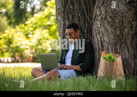 Guy typing on laptop sitting leaning on tree Stock Photo