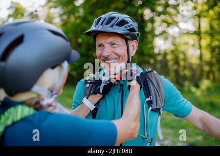 Active senior couple riding bicycles at summer park, man is putting on helmet, healthy lifestyle concept. Stock Photo