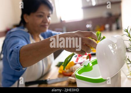 Biracial mature woman throwing vegetables waste in recycling bin while cooking food in kitchen Stock Photo