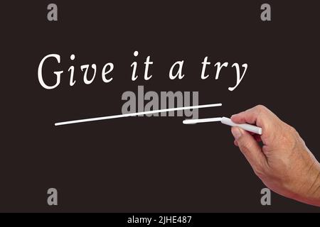 Male hand writes in white chalk pencil the word Give It A Try on a black background. Encouragement phrases and business concept. Stock Photo