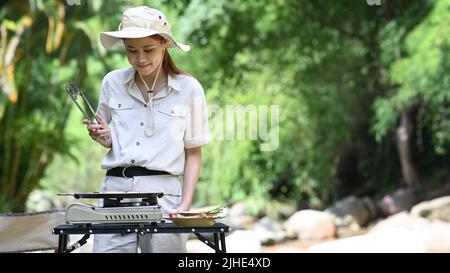 Young woman standing near creek and barbecuing meat on a portable gas stove. Travel, adventure on nature and vacation concept Stock Photo