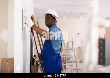 Painter in blue overalls leveling plaster on the wall with spatula Stock Photo
