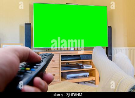 couch potato Man with feet up watching a large flat green screen tv remote control in hand changing channels Stock Photo