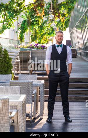 A young hipster-looking man in a suit with a bow tie, a manager in a summer street cafe, waits for visitors before opening Stock Photo