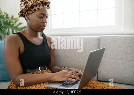 Close up Beautiful traditional young gen z African woman sitting on sofa at home, typing on laptop, smiling, shot with copy space Stock Photo