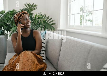 Beautiful young African woman sitting on sofa holding smart phone, smiling and laughing, shot with copy space Stock Photo