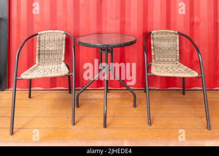 two wicker chairs and glass table near red wall of roadside cafe, empty Stock Photo