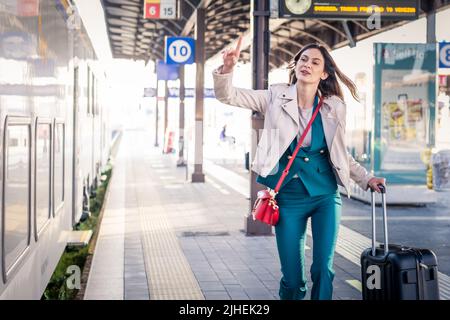 Beautiful girl running and chasing the leaving train in station. Waving hand and rushing to get on - Young business woman with suitcase running Stock Photo