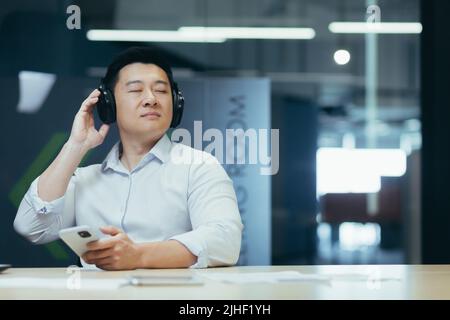 Asian boss business owner relaxing in office, listening relaxing and relaxing music, man sitting at desk, using big headphones and online listening app on phone Stock Photo