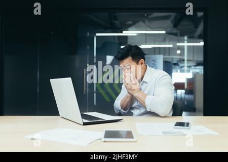 Sick man in office coughs and has runny nose, Asian businessman with cold works in office on laptop Stock Photo