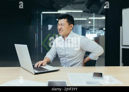 Asian businessman working in office, having severe back pain, overtired worker working with laptop. Stock Photo