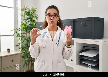 Young hispanic woman holding vaccine and piggy bank puffing cheeks with funny face. mouth inflated with air, catching air. Stock Photo