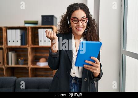 Young hispanic woman using touchpad wearing headphones screaming proud, celebrating victory and success very excited with raised arm Stock Photo