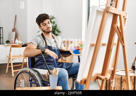 Young hispanic man sitting on wheelchair painting at art studio looking sleepy and tired, exhausted for fatigue and hangover, lazy eyes in the morning Stock Photo