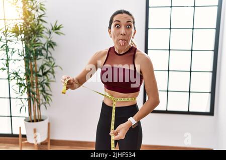 Young hispanic woman wearing sportswear using tape measure at sport center sticking tongue out happy with funny expression. Stock Photo