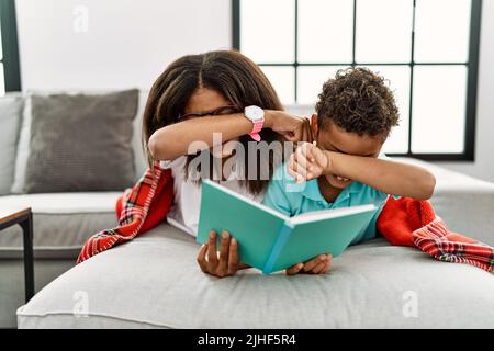 Two siblings lying on the sofa reading a book smiling cheerful playing peek a boo with hands showing face. surprised and exited Stock Photo