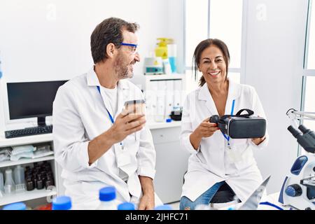 Middle age man and woman partners wearing scientist uniform drinking coffee and holding vr goggles at laboratory Stock Photo
