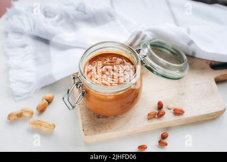 Peanut butter in glass jar, scattered peanuts on cutting board on white wooden table. Top view. Stock Photo