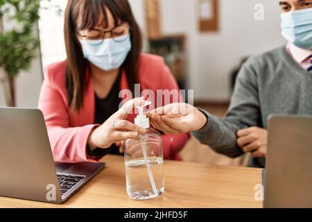 Two hispanic business workers wearing medical mask using sanitizer hands gel at the office. Stock Photo