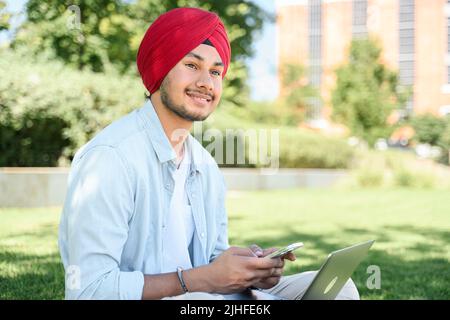 Smiling Indian male student in national turban sitting outdoors with laptop, using smartphone, texting online, chatting in social media network, using dating app. Side view Stock Photo
