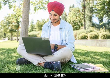 Serene Indian male student in traditional turban using laptop for online studying sitting outdoors on the lawn in campus area, preparing homework, researching topic, positive hindu guy typing Stock Photo