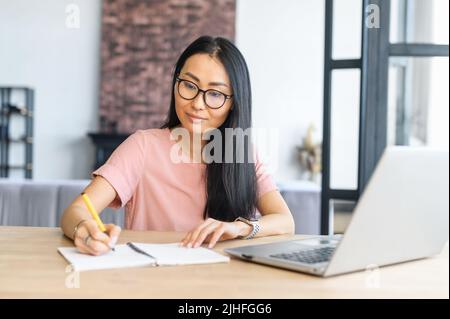 Mature asiatic woman receives a second education online, studies remotely, writes notes, looks well-groomed, copying information from internet site, using laptop while working on university essay Stock Photo