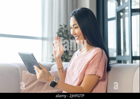 Young nice woman make video call using digital tablet while sitting on a sofa at home, waving her hand in greeting, chatting with relatives, friends, meeting on a dating site, pleasant conversation Stock Photo