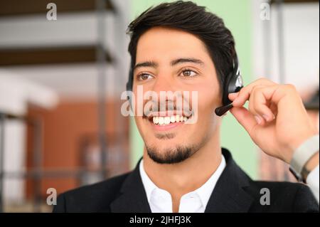 Smiling young latin male office employee wearing using headset and looking aside, positive man with the beard working in customer service department, making and receiving calls, sitting on workplace Stock Photo