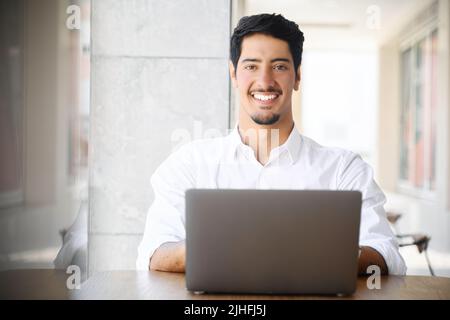 Front view of positive ethnic businessman in formal wear using laptop sitting outdoors, hispanic male employee looking at camera, cheerfully smiles, dark-haired handsome guy in white shirt with laptop Stock Photo