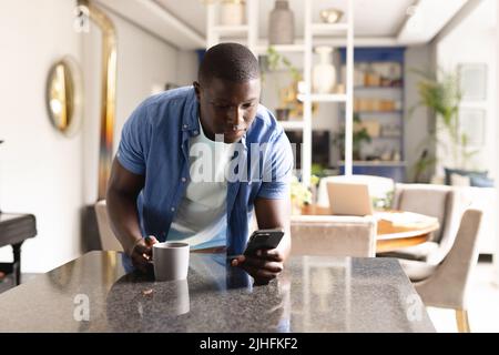 Vertical image of african american man drinking coffee and using smartphone Stock Photo