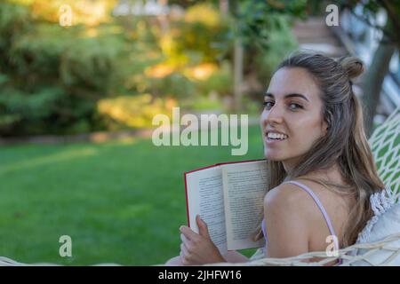 Young woman from the back looking to camera reading a book as leisure activity in the park of the city in spring Stock Photo