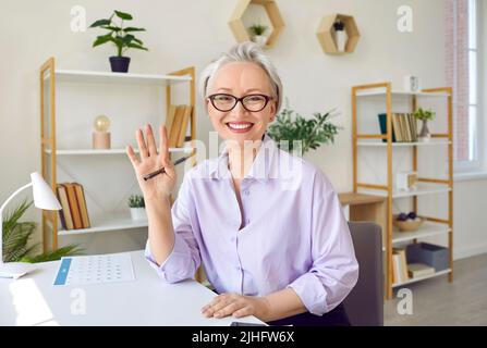 Happy mature female teacher waving hand during video conference calling in virtual webcam chat. Stock Photo