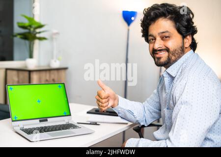 Happy young man with green screen laptop showing thumbs up by looking camera at office - concept of career promotion, advertisement and employment Stock Photo