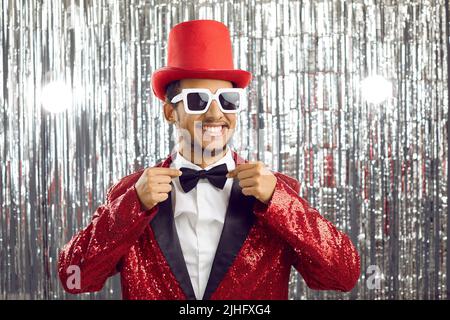 Funny guy in red sequin party suit, top hat and sunglasses smiles and fixes his bowtie Stock Photo