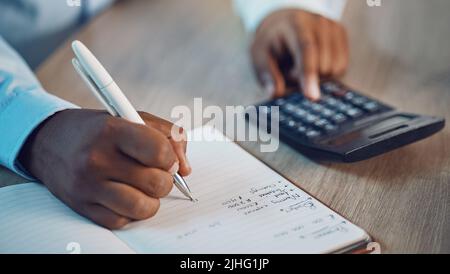 Closeup hands of african man writing in his diary while using a calculator. African american business man calculating figures while working late at Stock Photo