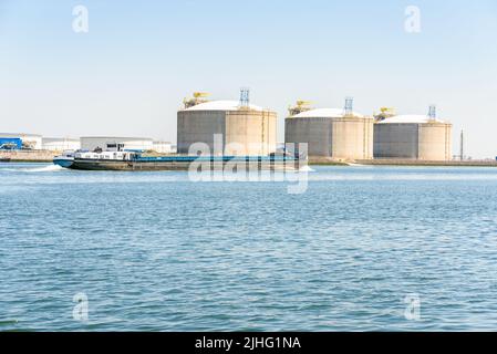 Bulk carrier barge in harbour with large concrete tanks for storage of liquefied natural gas in background Stock Photo