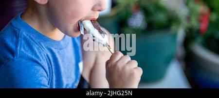 young boy eating an ice-cream bar with a chocolate coating outside on a hot day.  Stock Photo