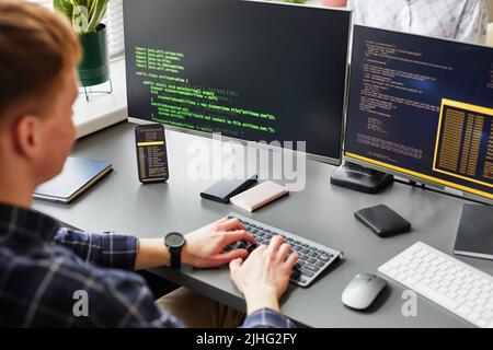 Rear view of young software developer writing codes of new software sitting at his workplace with computer monitors Stock Photo