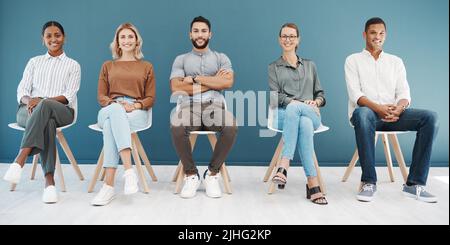 Portrait of a diverse group of confident business men and women sitting on chairs in waiting room against a blue background. Happy hopeful candidates Stock Photo